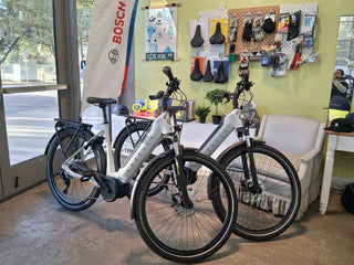 Two white Bosch electric bicycles displayed indoors in a shop setting, with a sofa, wall-mounted accessories, and a window visible in the background.